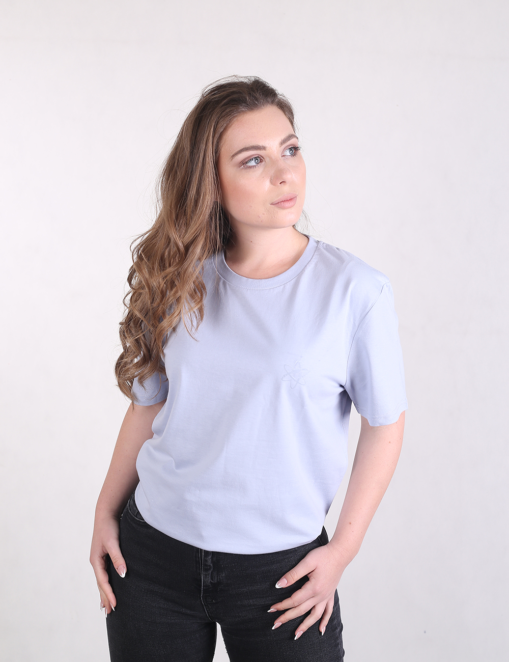 A woman with long wavy brown hair looking off to the side, wearing a light blue crew-neck t-shirt and black jeans against a plain white background.