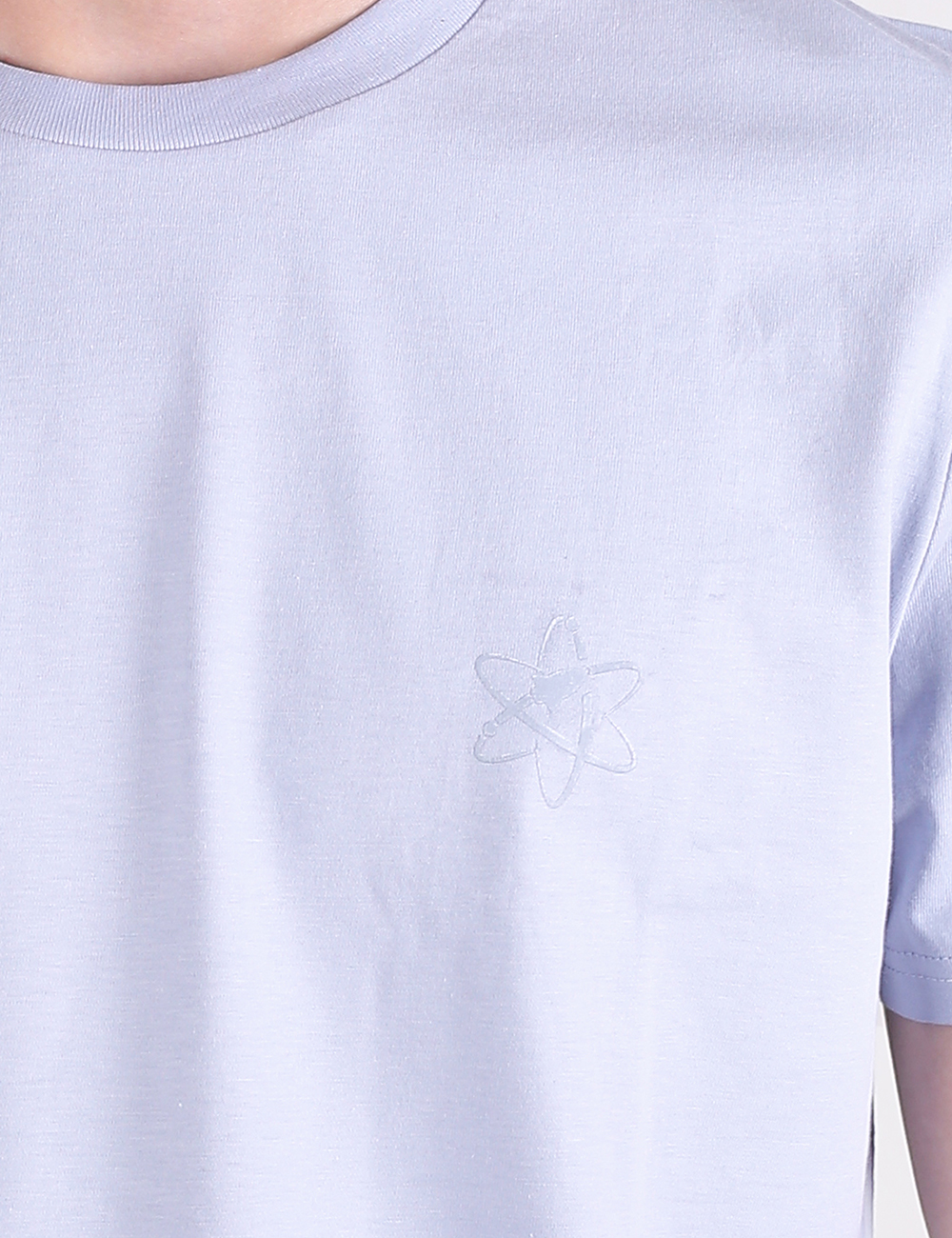 A close-up shot of a light blue t-shirt featuring a subtle, tonal atom symbol embroidered or printed on the left side of the chest.