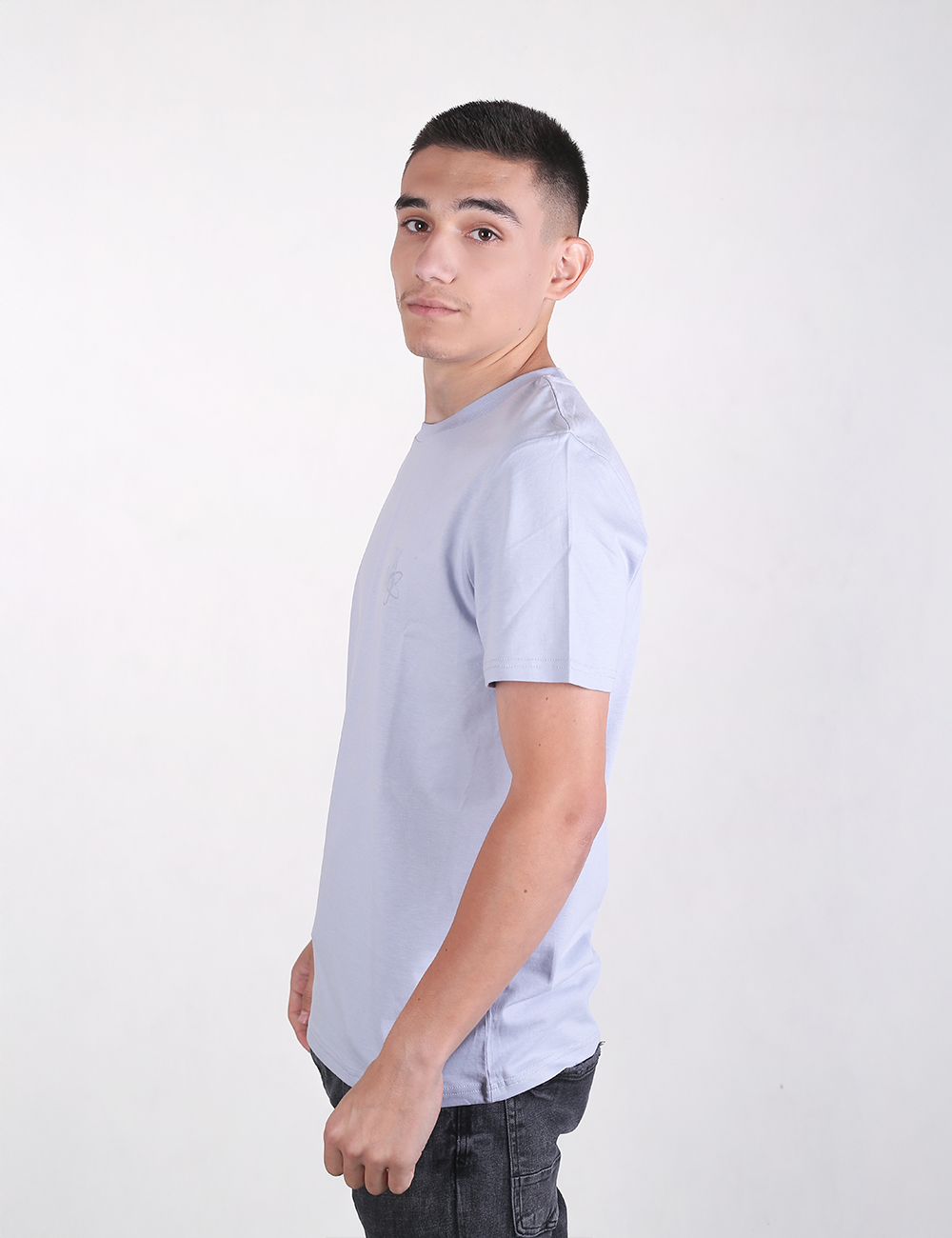 A young man with a short haircut stands in a side-profile view against a solid white background, wearing a light blue crew-neck T-shirt and dark jeans.