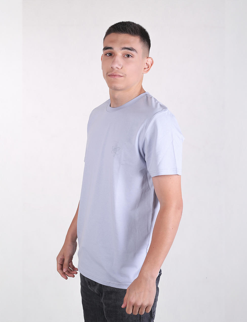 A young man with short dark hair standing in profile against a plain white background, wearing a light blue short-sleeved crew-neck t-shirt and dark jeans.
