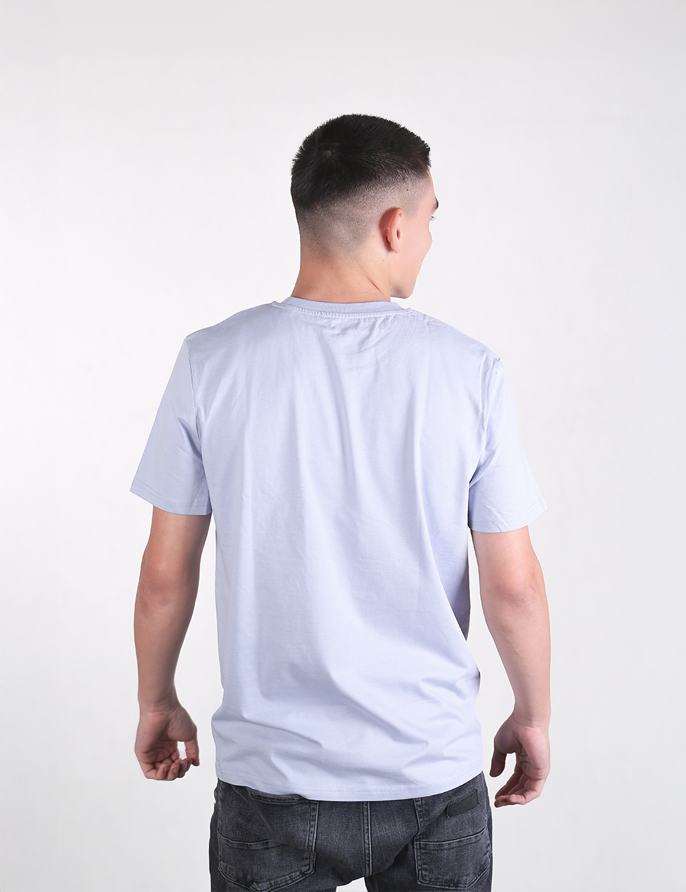 A young man with a short fade haircut is shown from the back, wearing a light blue, solid-colored crewneck t-shirt and dark gray jeans.