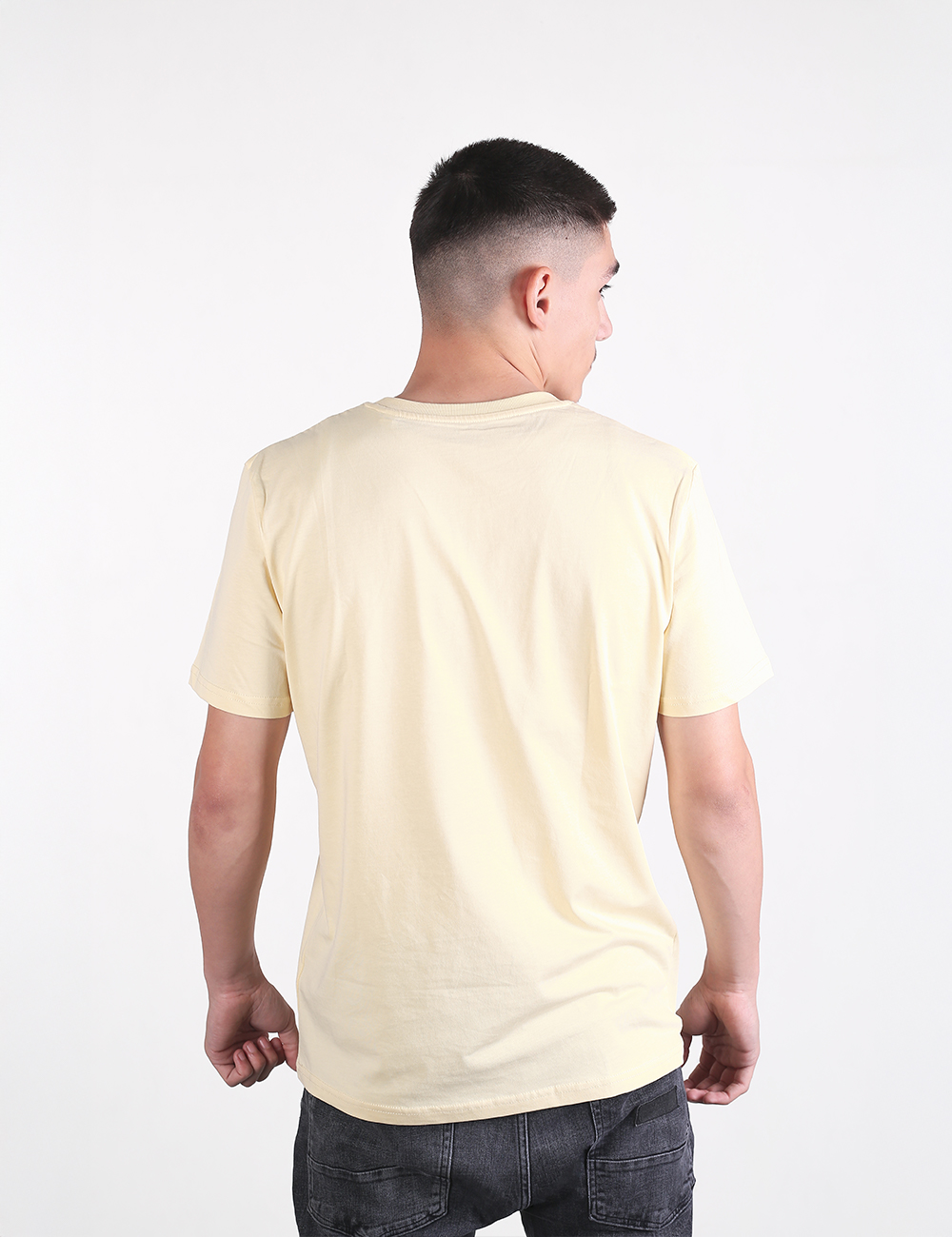 A man seen from behind is wearing a short-sleeved, pale yellow crewneck t-shirt and dark blue or black jeans. He has a fade haircut, and his arms are down at his sides with his fists lightly clenched. The background is solid white.