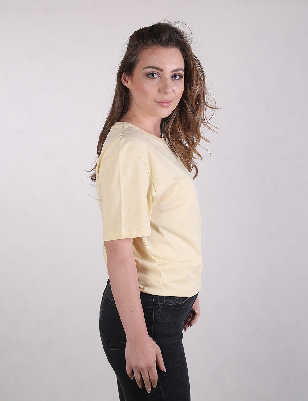 A woman with long, wavy brown hair styled over one shoulder is shown from a side profile, looking toward the camera. She is wearing a light yellow, short-sleeved crew-neck t-shirt and dark charcoal  jeans. 