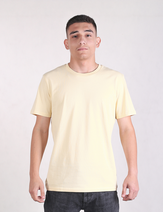 A young man with short dark hair stands facing forward, wearing a plain, light yellow crew neck t-shirt and dark gray pants. He has a neutral expression and is positioned against a solid, light gray background.