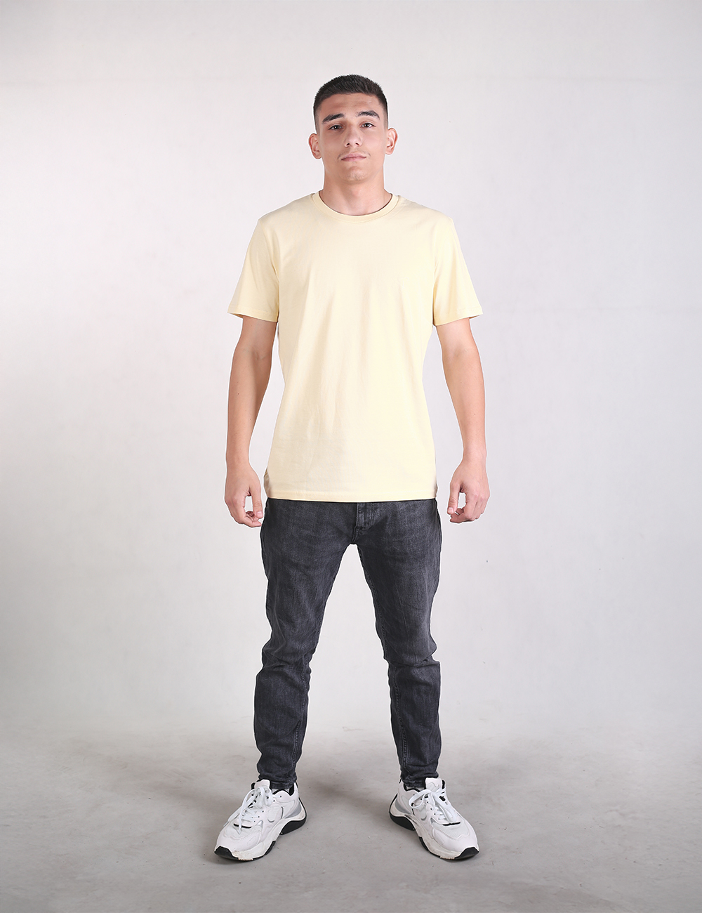A young man stands facing forward against a plain light gray background, wearing a light yellow crew-neck t-shirt, dark gray slim-fit jeans, and white sneakers. He is positioned in the center of the frame with a neutral expression, arms down at his sides, and feet set slightly apart.