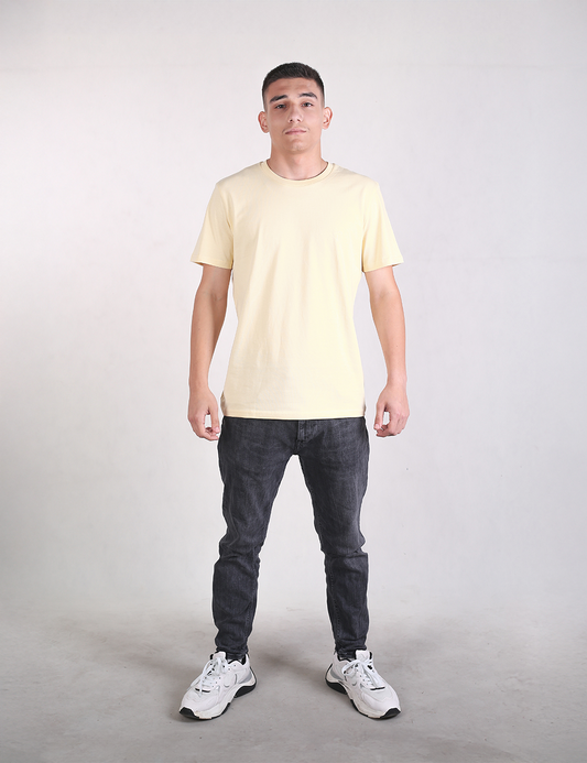 A young man stands facing forward against a plain light gray background, wearing a light yellow crew-neck t-shirt, dark gray slim-fit jeans, and white sneakers. He is positioned in the center of the frame with a neutral expression, arms down at his sides, and feet set slightly apart.