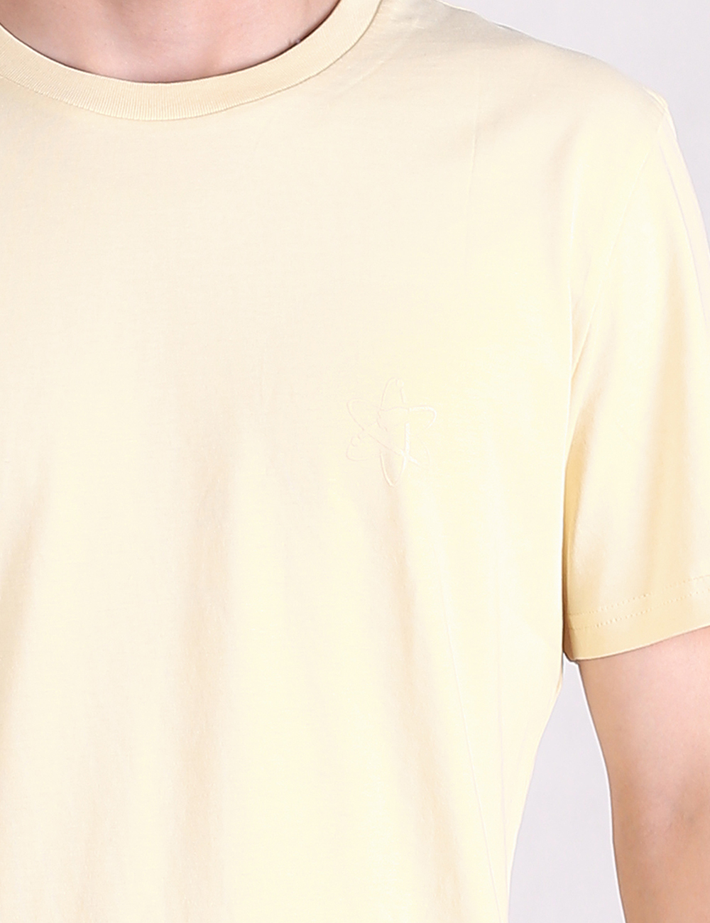 A close-up shot of a person wearing a light yellow, crew-neck, short-sleeved T-shirt. The image focuses on the upper torso, chest, and right shoulder, showcasing the smooth, plain fabric of the garment.