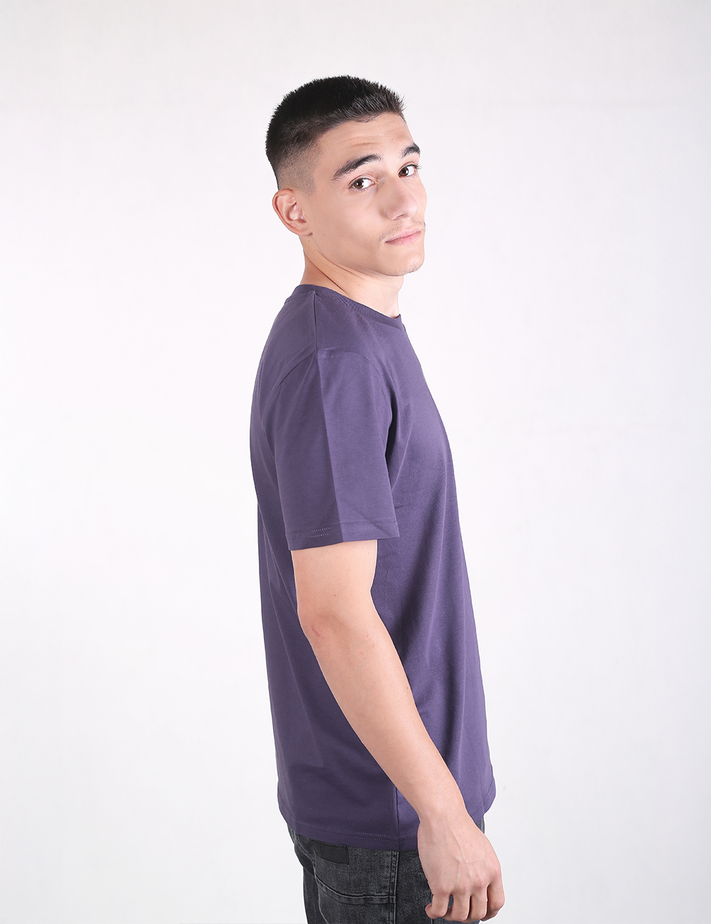 A young man with short dark hair, shaved on the sides and back, stands looking back over his right shoulder against a plain white background. He is wearing a dark purple crewneck short-sleeve t-shirt and dark jeans, shown from the side in a waist-up profile view.