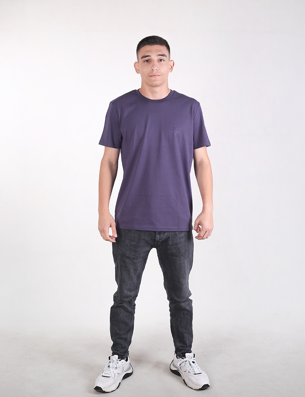 Full-length studio shot of a young man standing against a solid off-white background, facing forward. He has short dark hair and a neutral expression.He is wearing a solid dark purple crewneck t-shirt, slim-fit dark gray jeans, and white athletic sneakers with black accents. He is standing with his feet apart and his arms relaxed at his sides.