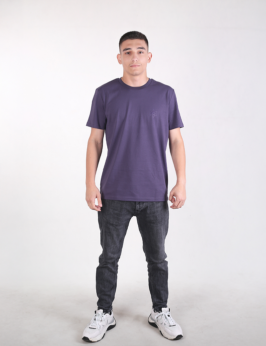Full-length studio shot of a young man standing against a solid off-white background, facing forward. He has short dark hair and a neutral expression.He is wearing a solid dark purple crewneck t-shirt, slim-fit dark gray jeans, and white athletic sneakers with black accents. He is standing with his feet apart and his arms relaxed at his sides.