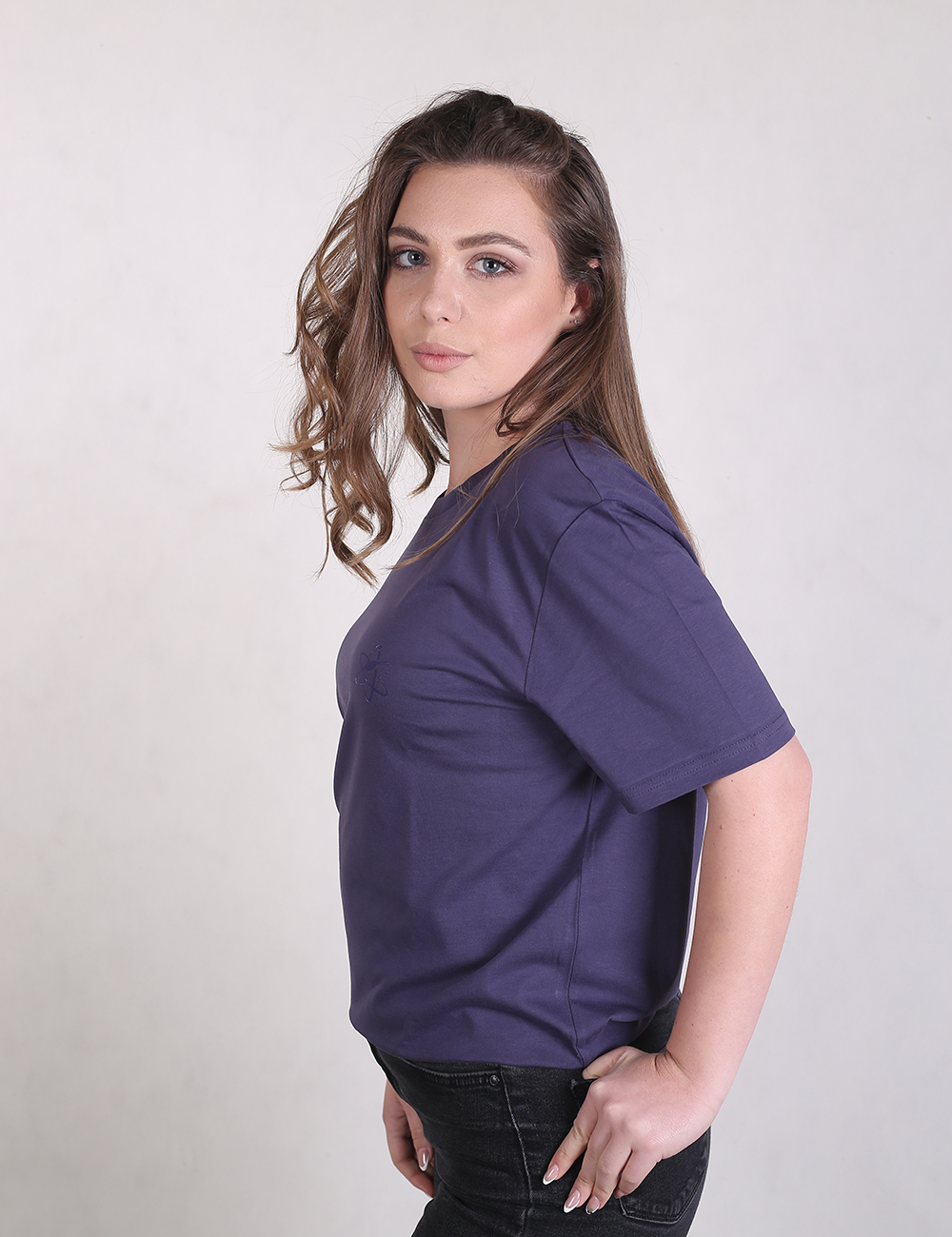 A young woman with long, light brown hair styled to one side is posing in a side profile view against a plain, light grey background. She is wearing a dark purple crew-neck t-shirt with short sleeves and a relaxed fit, paired with black or dark denim jeans. She has her left hand resting on her hip, slightly tucked into her pocket, and is looking directly at the camera.