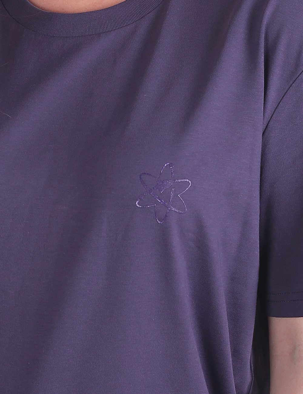 A close-up shot focuses on the left chest area of a loose-fitting, purple t-shirt. The crewneck collar and part of the right sleeve are visible. Embroidered on the chest is a small, subtle atom symbol in a matching purple thread.