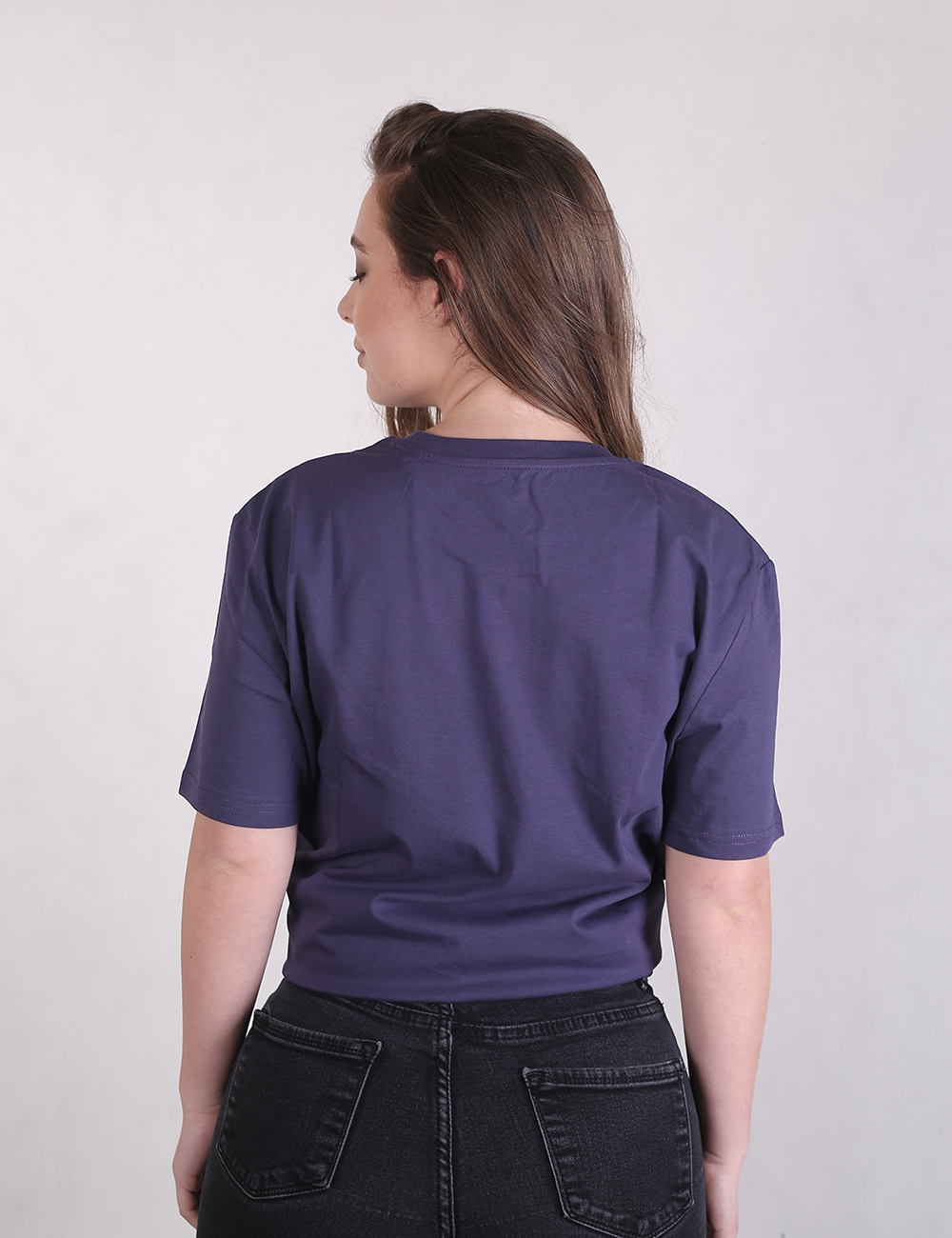 A woman with long brown hair stands facing away from the camera, showcasing the back of a short-sleeved, dark purple t-shirt and black denim jeans.