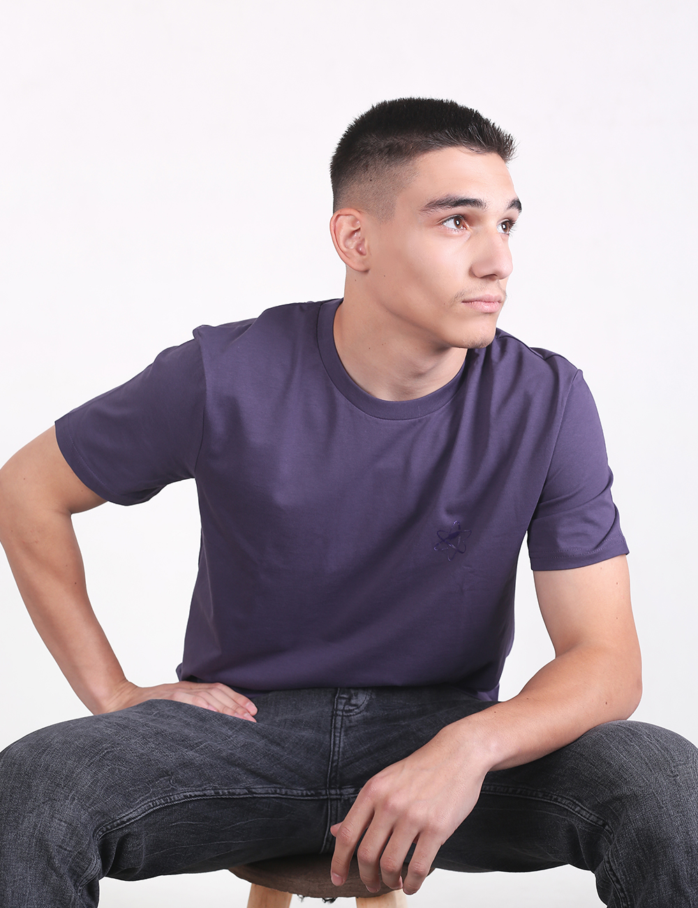 A medium shot of a young man with dark hair in a short buzz cut fade, looking off to his right against a plain white background. He is wearing a solid dark purple or indigo short-sleeve crewneck t-shirt and dark grey, acid-wash jeans. He is seated with one hand resting on his hip and the other on his leg.