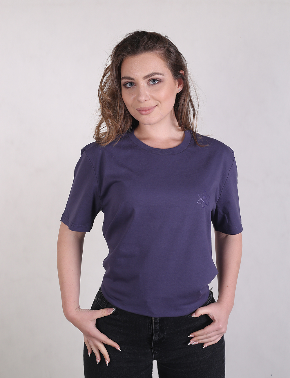 A woman with long wavy brown hair, wearing a dark purple or navy blue crew neck short-sleeve t-shirt and black jeans, posing against a light grey plain background with her hands on her waist.