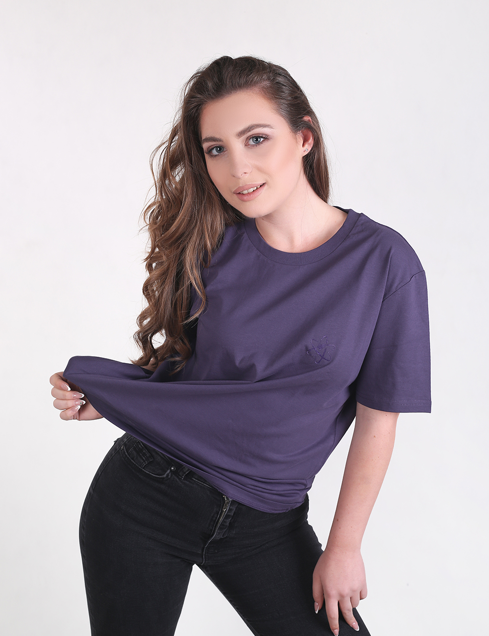 A young woman with long, wavy brown hair styled over one shoulder smiles at the camera, wearing an oversized, short-sleeved dark purple t-shirt and slim-fit black jeans.