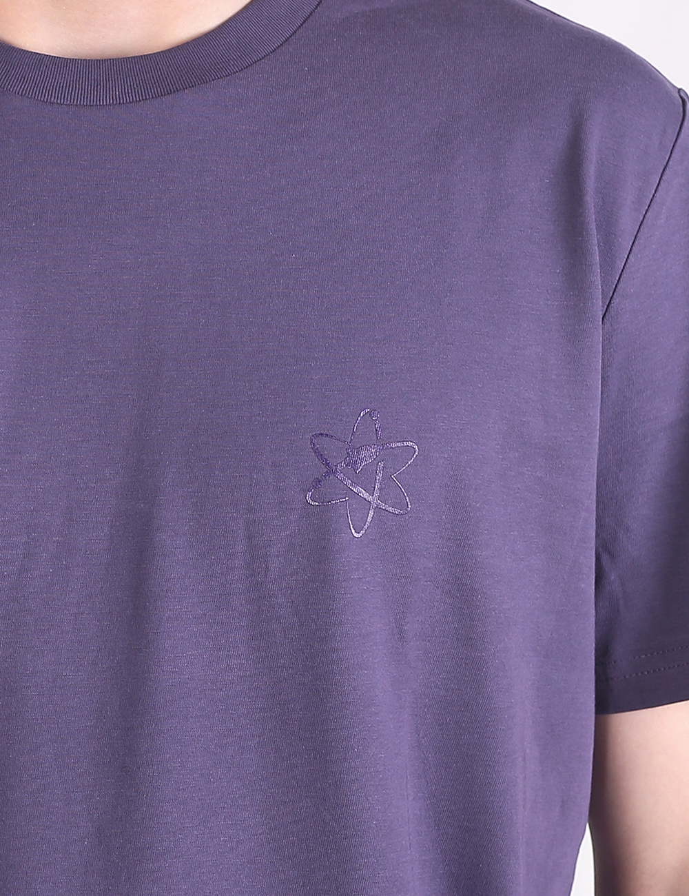 A close-up shot of a person wearing a dark lavender or purple crewneck t-shirt. The fabric is smooth cotton, and the left chest features a subtle, tone-on-tone embroidered logo of an atomic symbol or star outline.