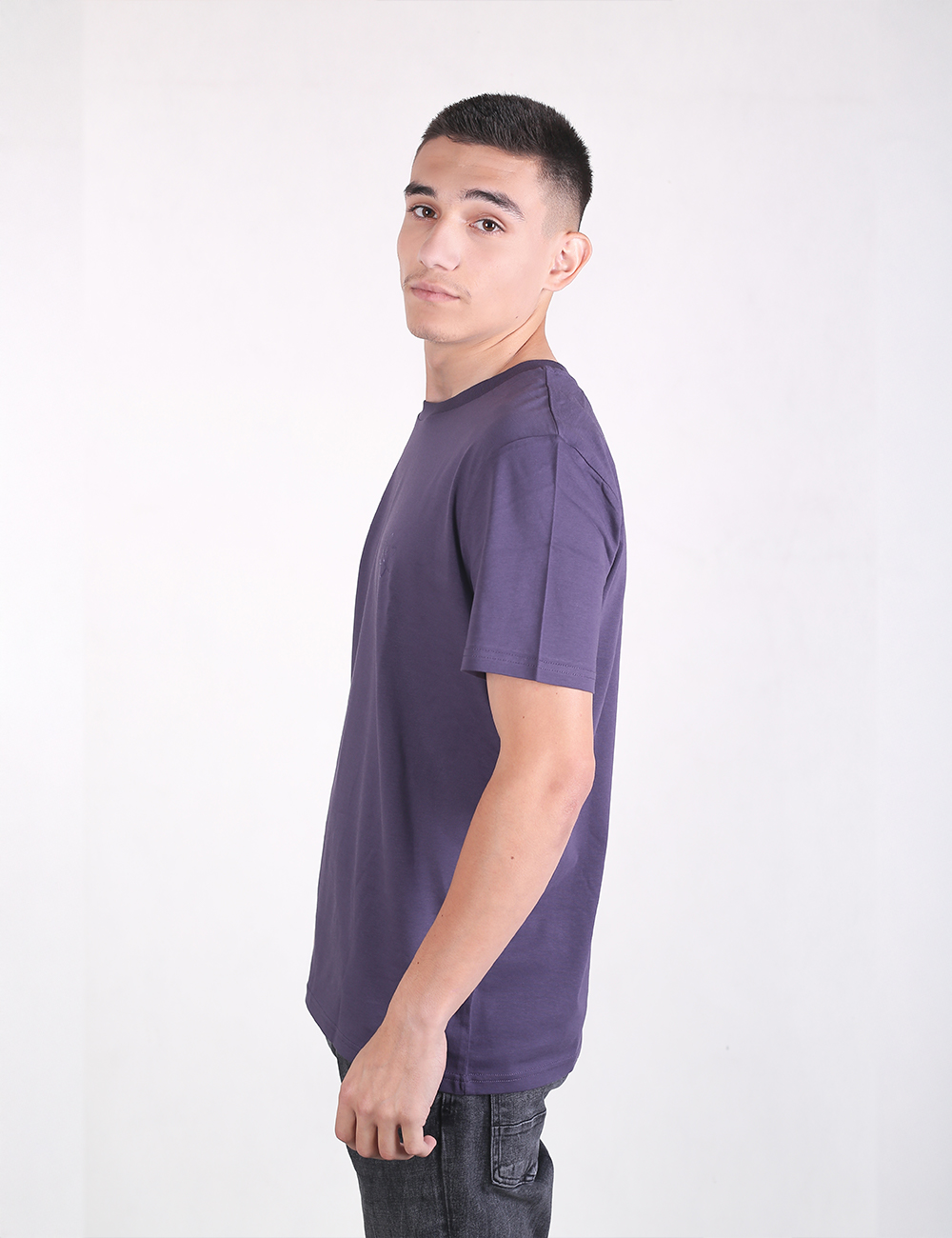 A young man with short dark hair standing in profile against a plain white background, wearing a dark purple crew-neck short-sleeve t-shirt and dark jeans.