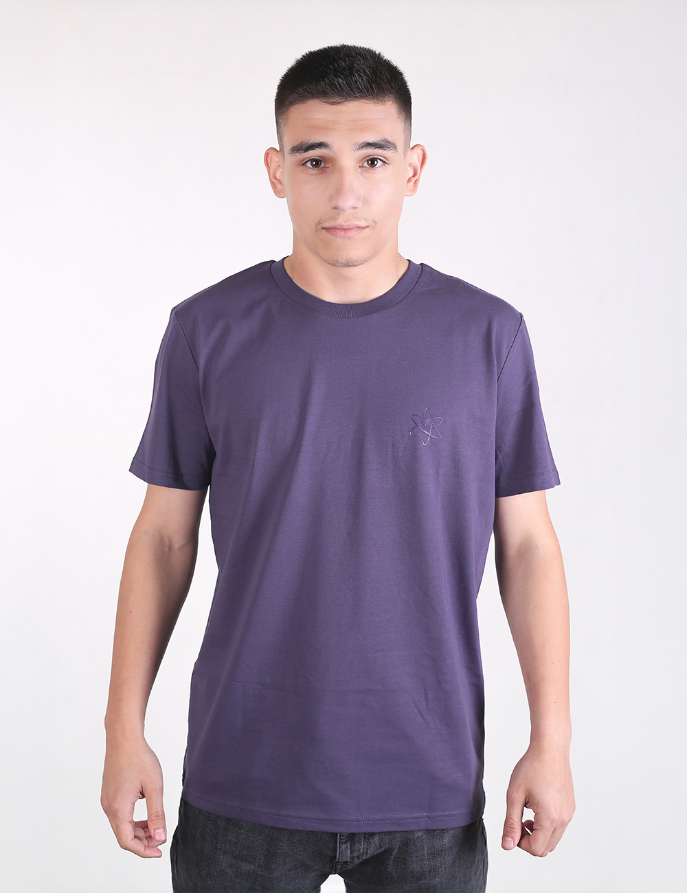A young man with short dark hair standing straight against a plain light gray background, wearing a casual, solid indigo-purple organic cotton t-shirt with a classic crew neck and short sleeves. The shirt features a subtle, tone-on-tone embroidered logo on the left chest.