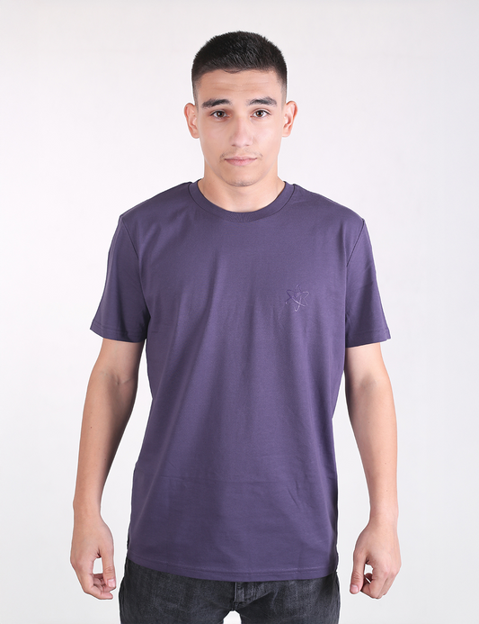 A young man with short dark hair standing straight against a plain light gray background, wearing a casual, solid indigo-purple organic cotton t-shirt with a classic crew neck and short sleeves. The shirt features a subtle, tone-on-tone embroidered logo on the left chest.