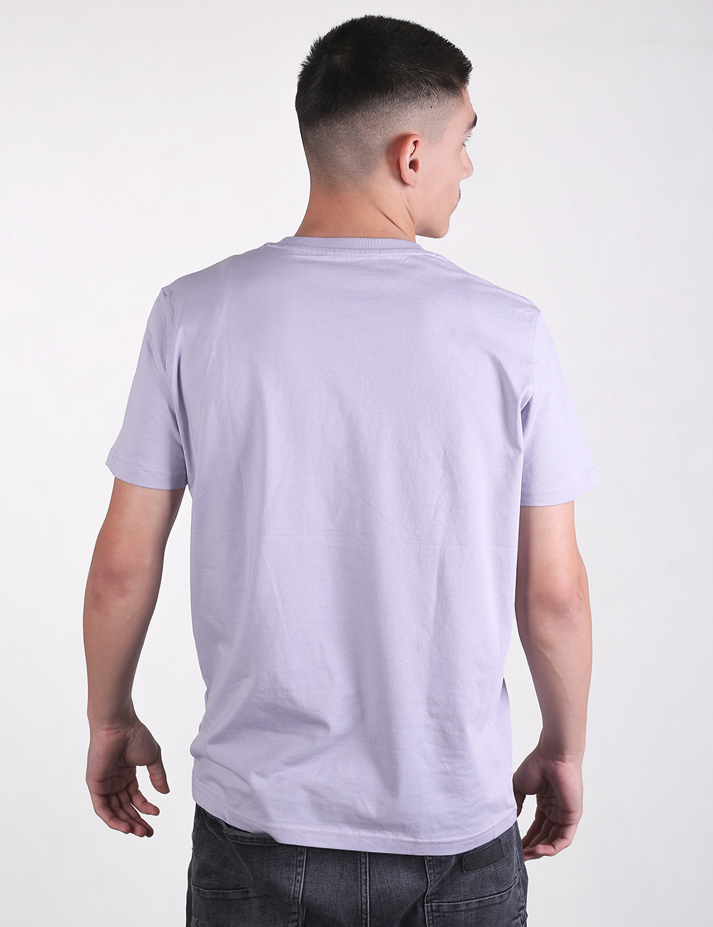 Rear-facing view of a young man with a short haircut featuring faded sides, wearing a plain, light lavender short-sleeve crewneck t-shirt and dark grey or black jeans, set against a solid, light grey background.