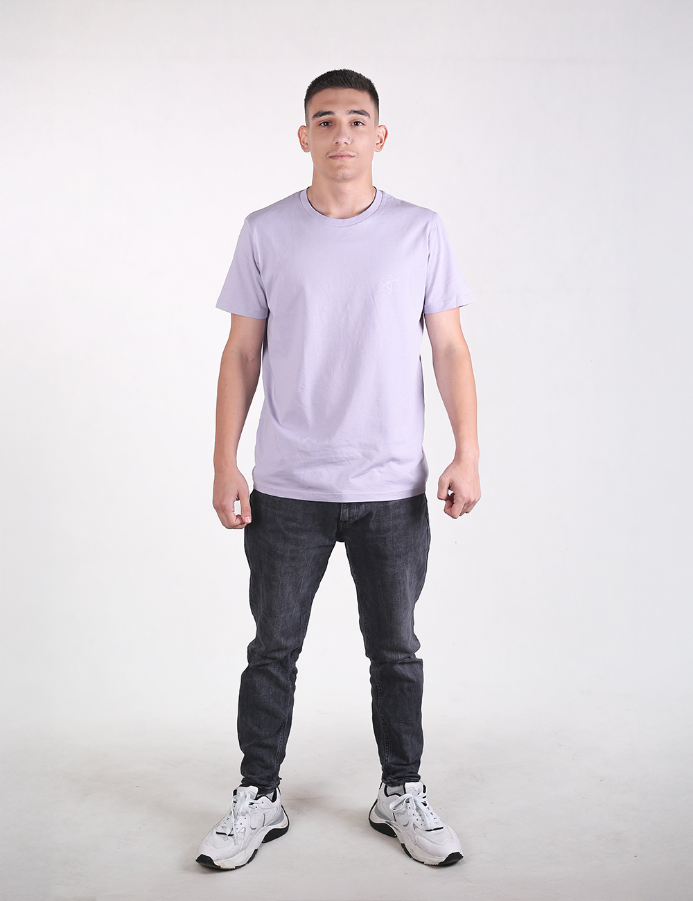 A man stands facing forward against a plain white background, wearing a light lavender crew-neck t-shirt, slim-fit dark grey jeans, and chunky white and grey athletic sneakers.