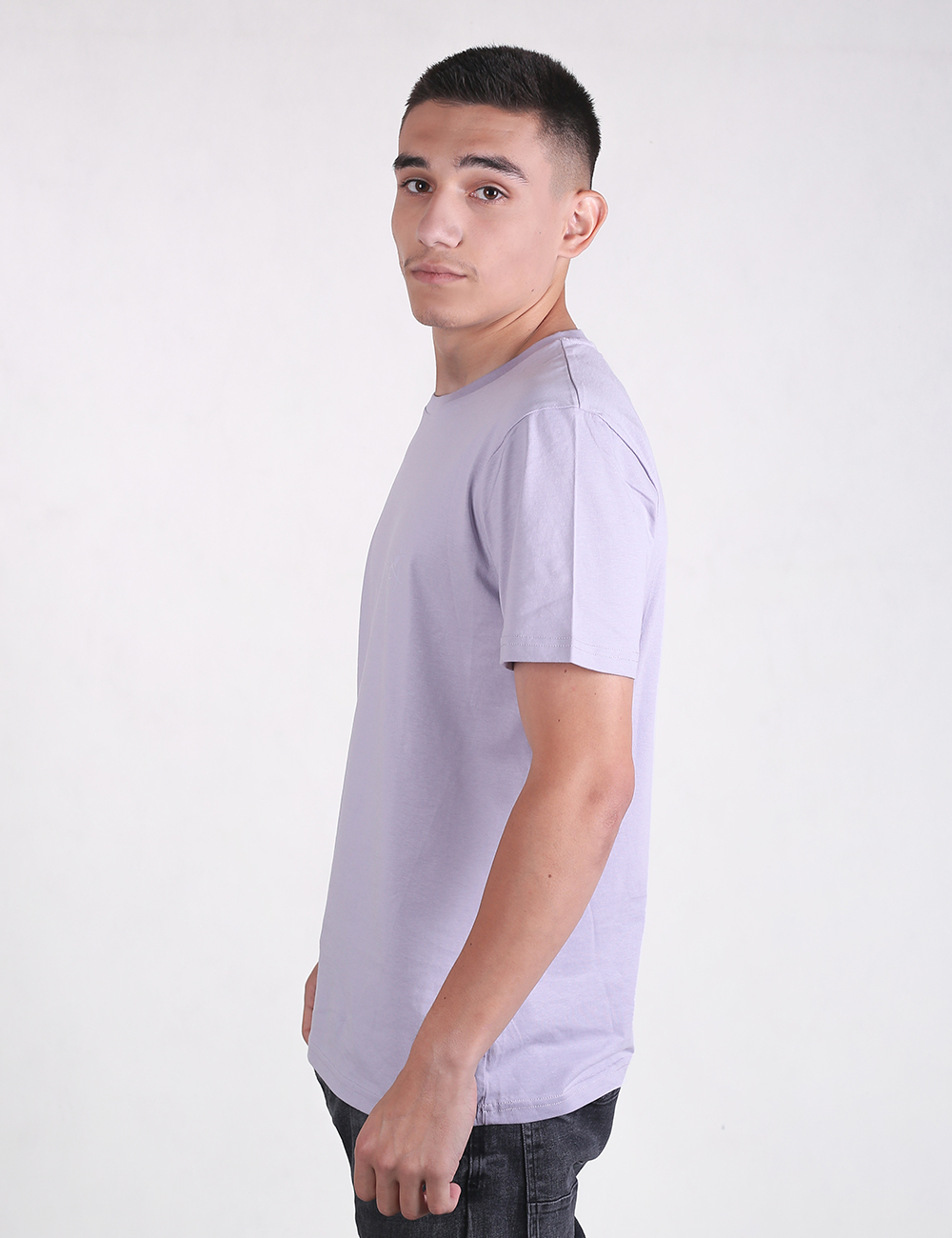 A young man with short dark hair standing in profile against a plain white background, wearing a light lavender or lilac short-sleeved crew neck T-shirt and dark jeans.