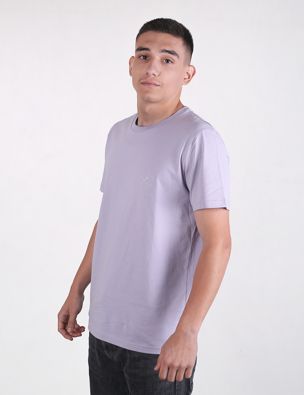 A young man  wearing a light lavender or light purple crewneck t-shirt and dark pants. The background is a solid, light grey color.