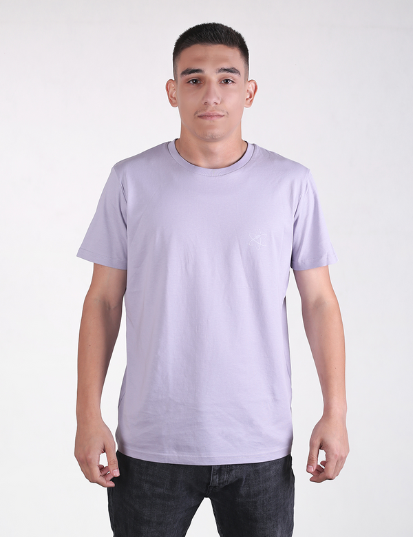 A man with short dark hair is standing against a plain white background, facing forward. He is wearing a light purple or lavender short-sleeve crew neck t-shirt and dark grey or black jeans.