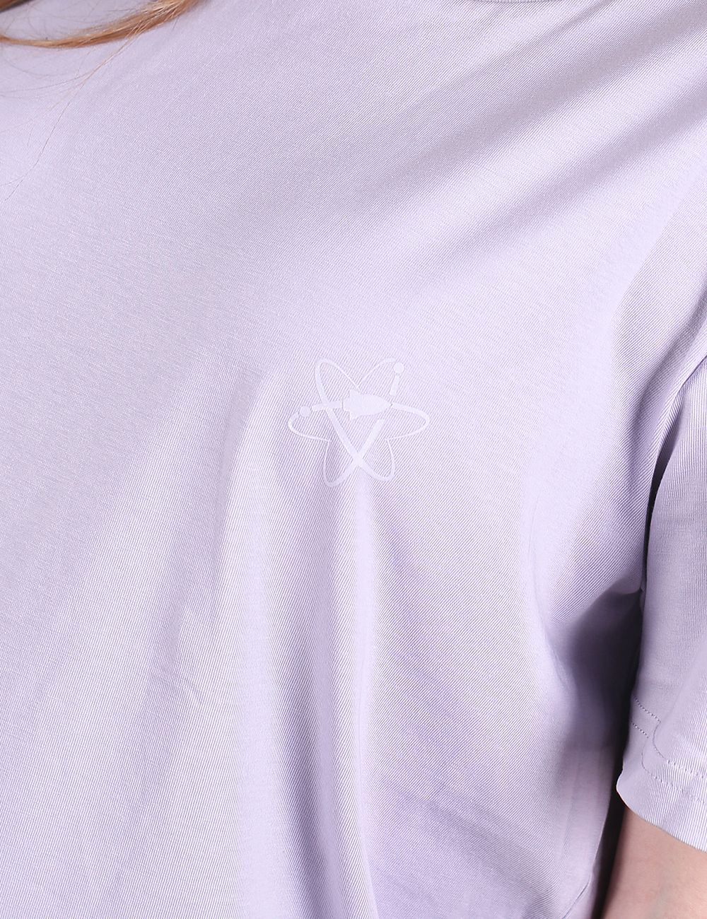 A close-up shot of a lilac or light purple t-shirt featuring a small, white embroidered or printed logo in the center of the chest. The minimalist logo resembles an atomic orbital structure or a stylized star shape. The fabric shows soft folds and wrinkles, with a hint of a sleeve visible on the right side.