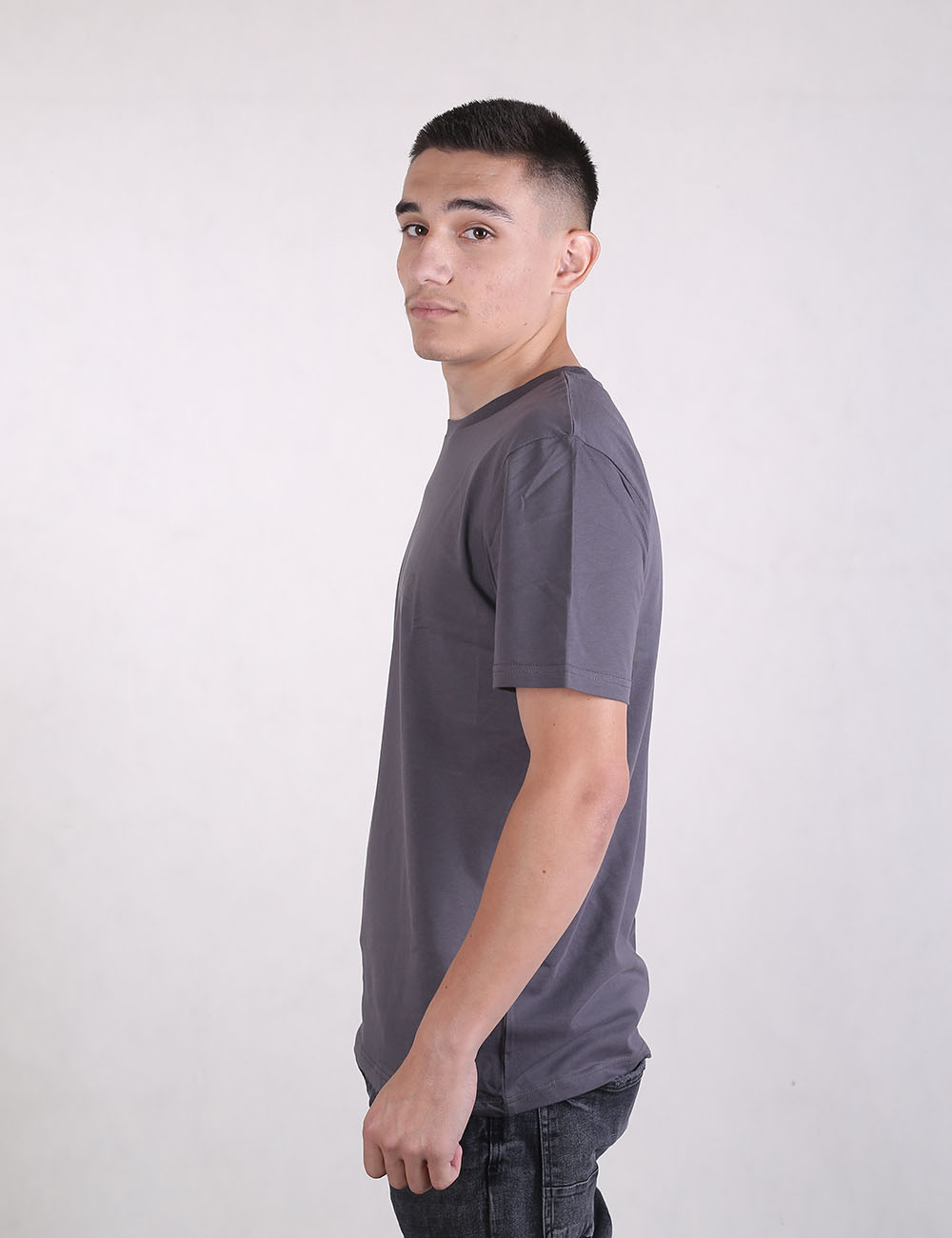 A young man with short dark hair stands in a side profile view against a plain, light grey background. He is wearing a short-sleeved, charcoal grey crew neck t-shirt and dark pants. He is looking back over his shoulder directly toward the camera with a neutral expression.