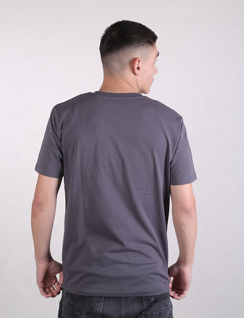 Rear view of a man with a short fade haircut, wearing a plain, solid charcoal grey t-shirt and black jeans against a light grey background.