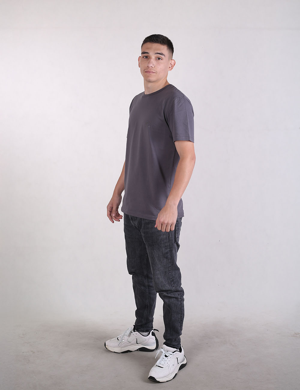 A full-body shot of a young man with short brown hair standing at a three-quarter angle against a plain light grey studio background. He wears a plain grey crewneck t-shirt, dark charcoal or faded black slim-fit jeans, and white athletic sneakers with black accents.
