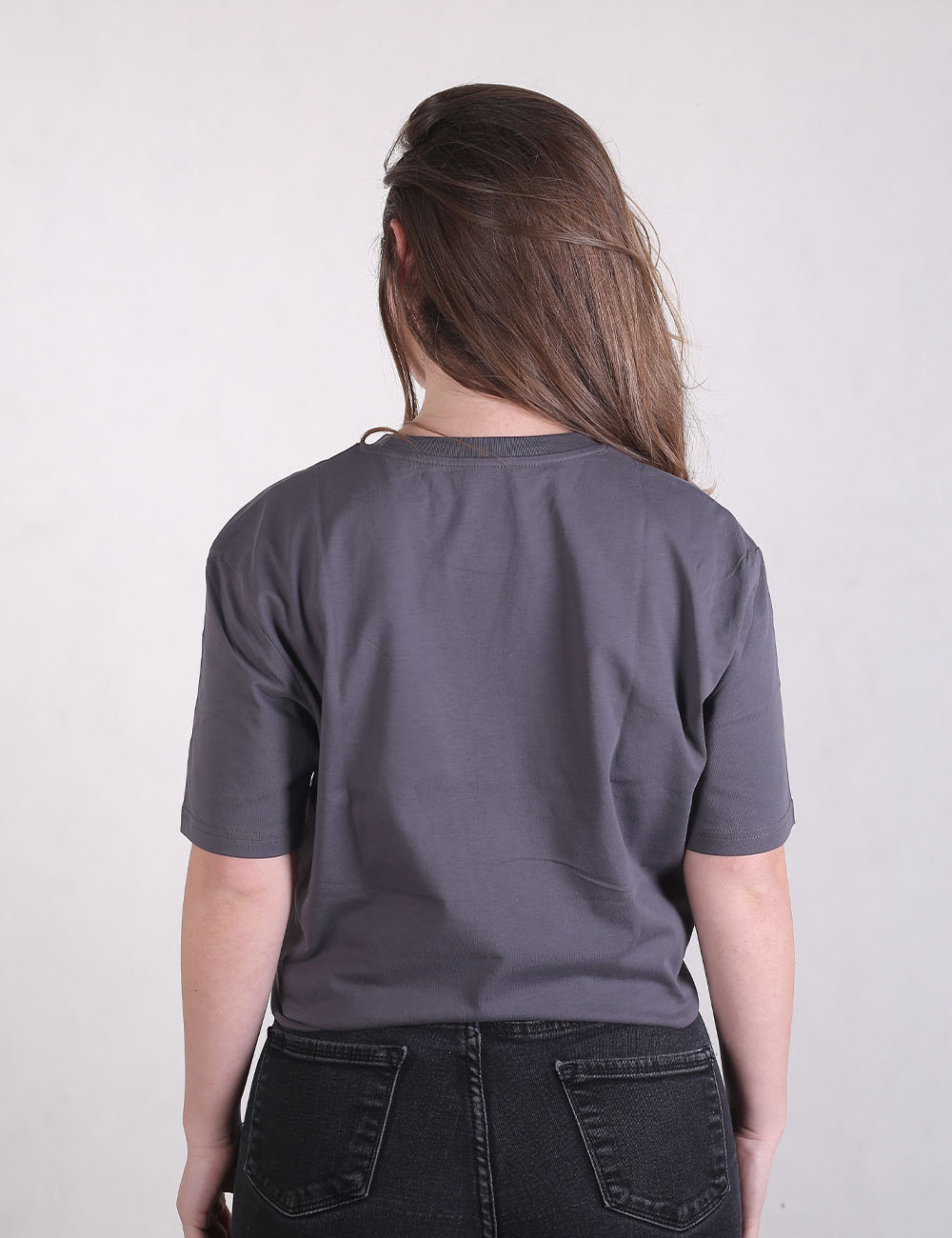 Rear view of a person with long brown hair wearing a loose-fitting charcoal grey t-shirt and dark denim jeans against a plain off-white background.
