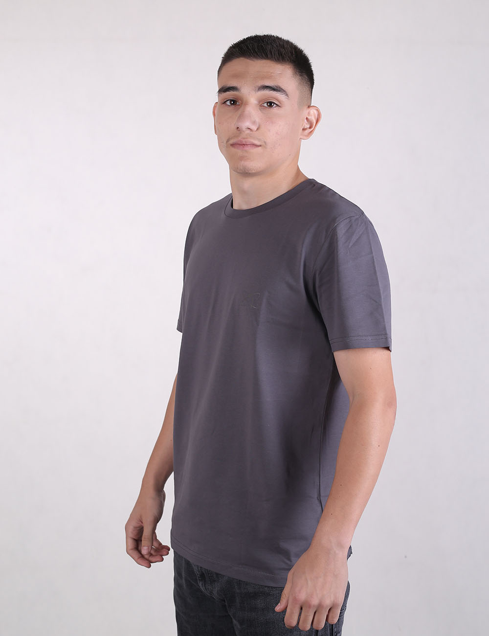 A young man standing in a side-profile view wearing a basic, charcoal grey short-sleeved crewneck t-shirt and dark jeans or trousers. He has short, dark cropped hair, a light complexion, and is looking directly at the camera with a neutral expression.