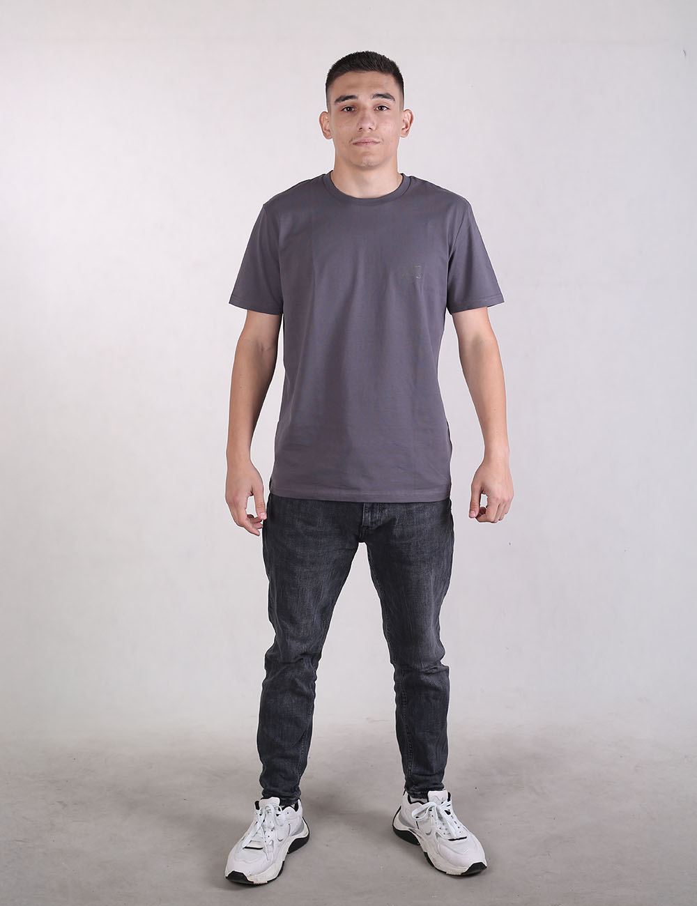 A man standing against a plain gray studio background, wearing a charcoal gray crew-neck t-shirt, dark gray or faded black jeans, and white athletic sneakers.