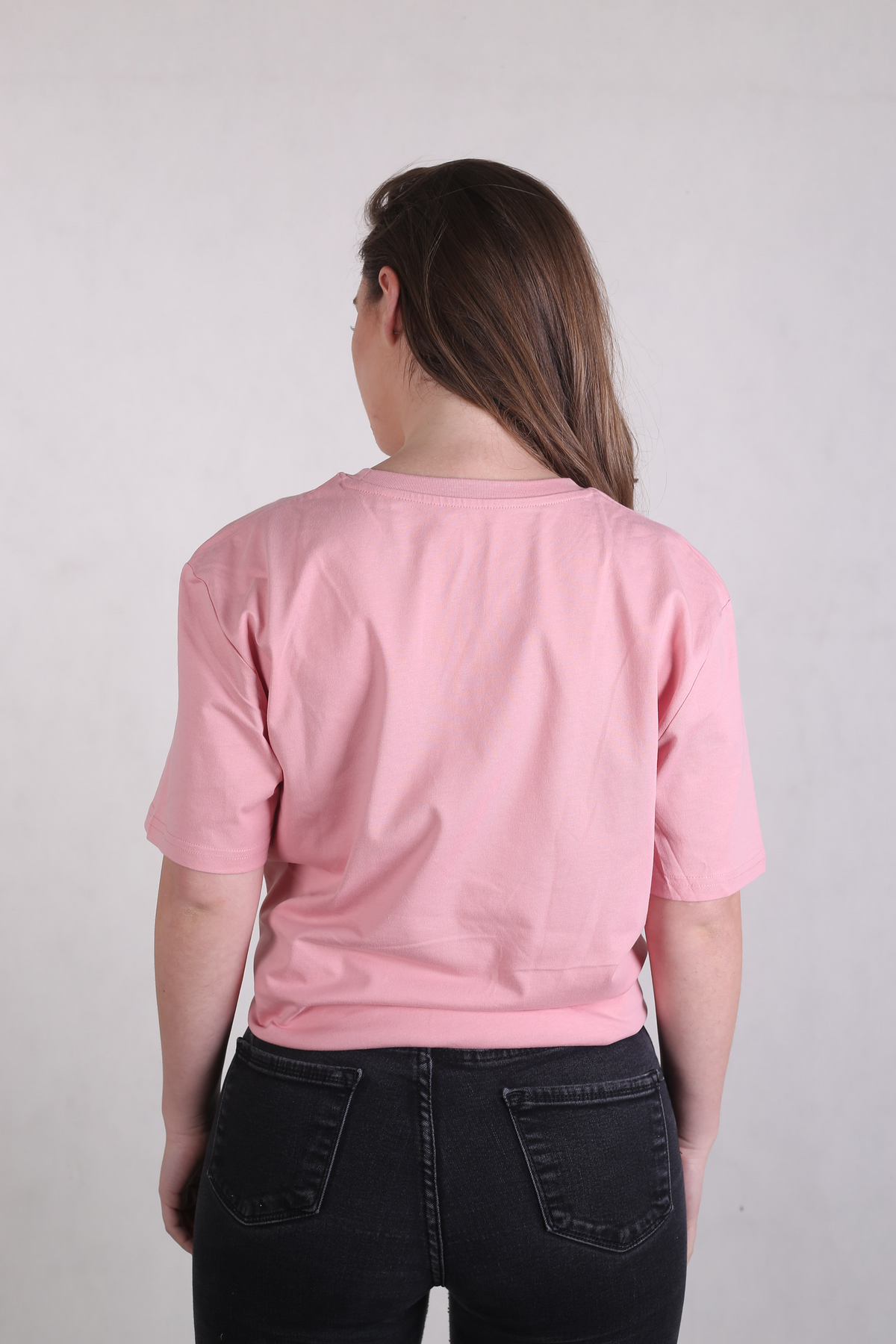 A woman viewed from behind wearing a light pink short-sleeve crewneck t-shirt and black jeans, standing against a plain light gray wall.
