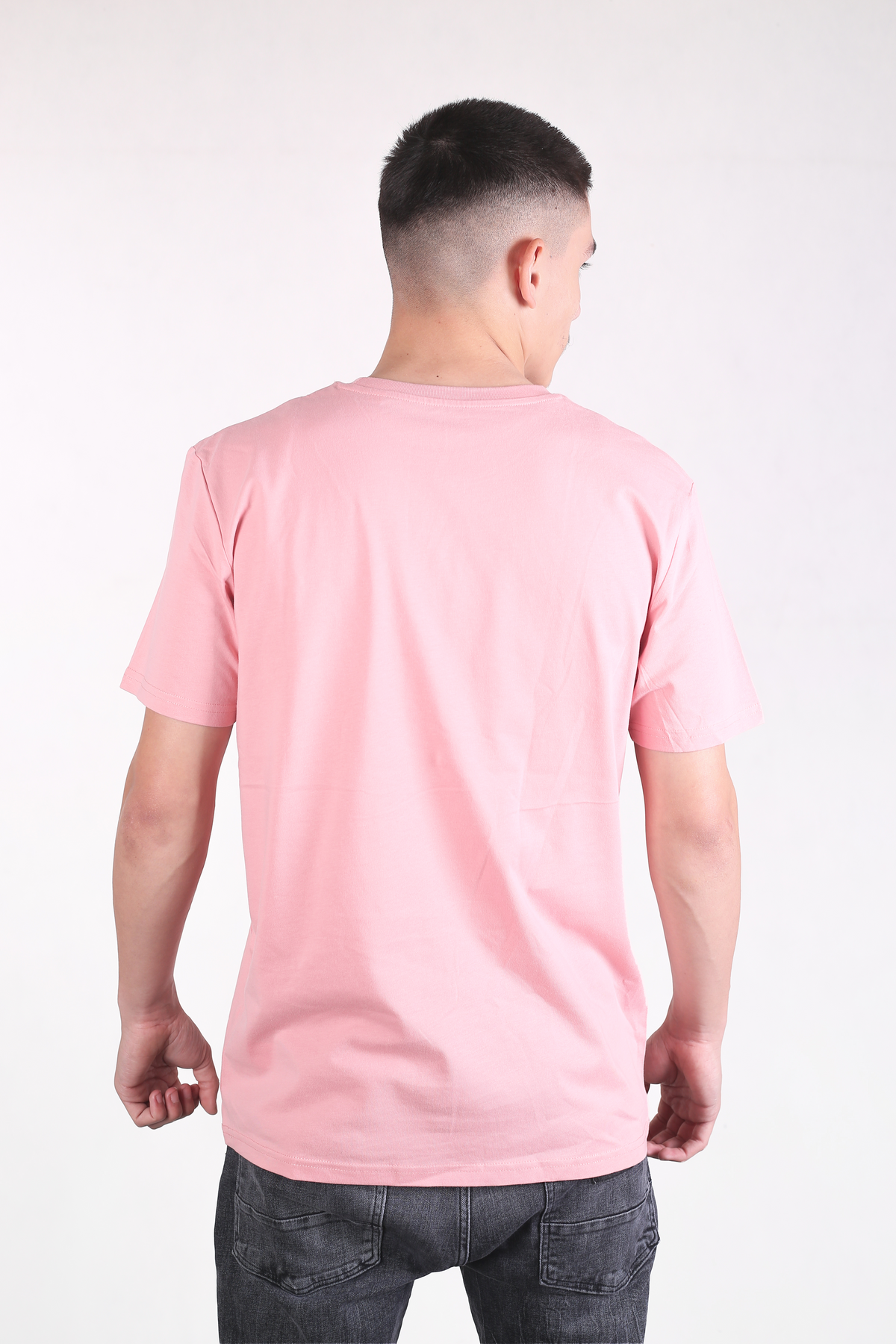 A man viewed from behind wearing a light pink short-sleeve crewneck t-shirt and dark gray jeans, standing against a plain white studio background.