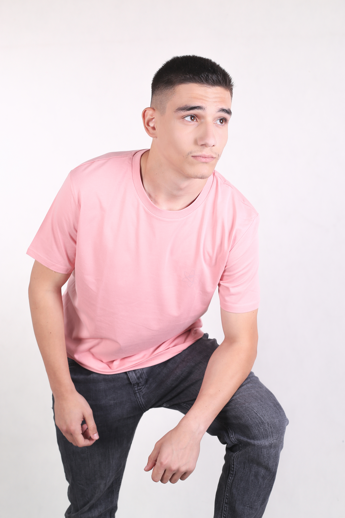 A man wearing a light pink crew-neck t-shirt and dark gray jeans, posing in a crouched position against a plain white studio background.