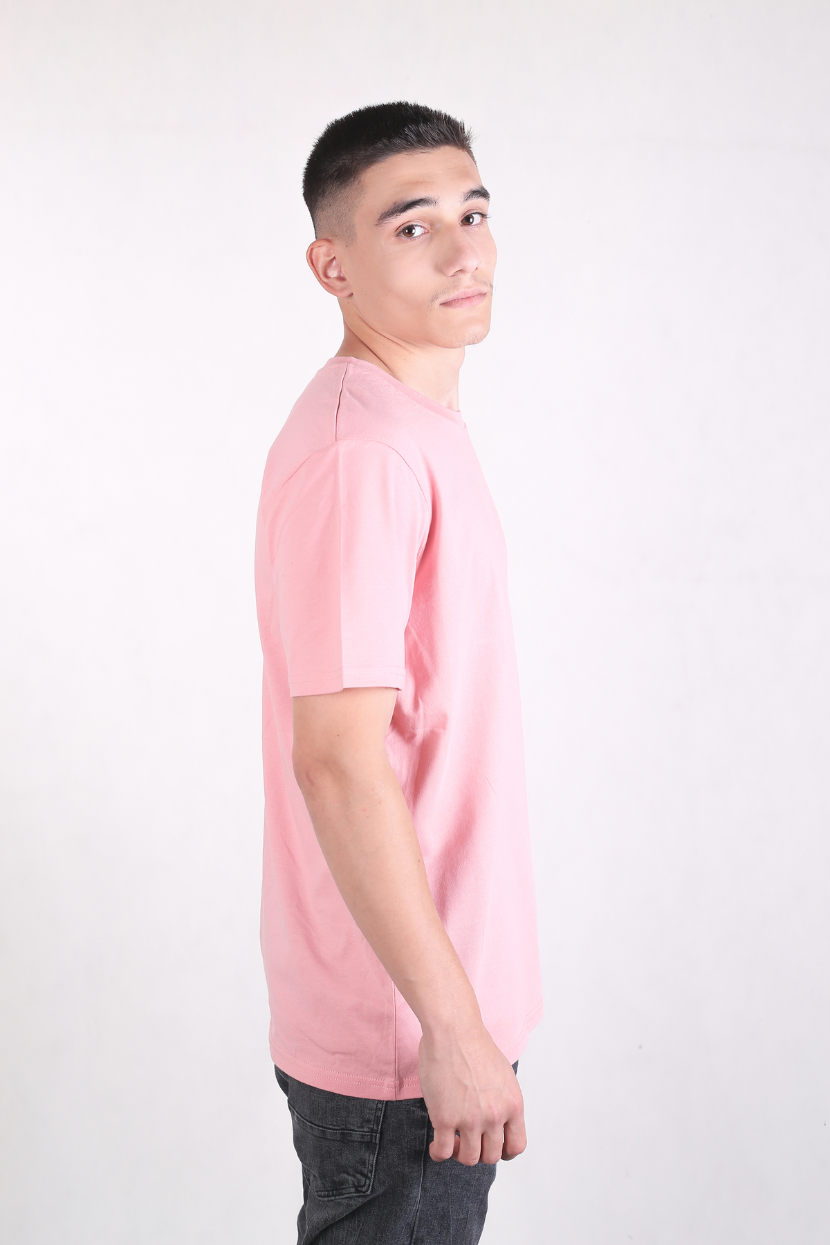 A man wearing a light pink crew-neck t-shirt and dark gray jeans, standing in profile against a plain white studio background.