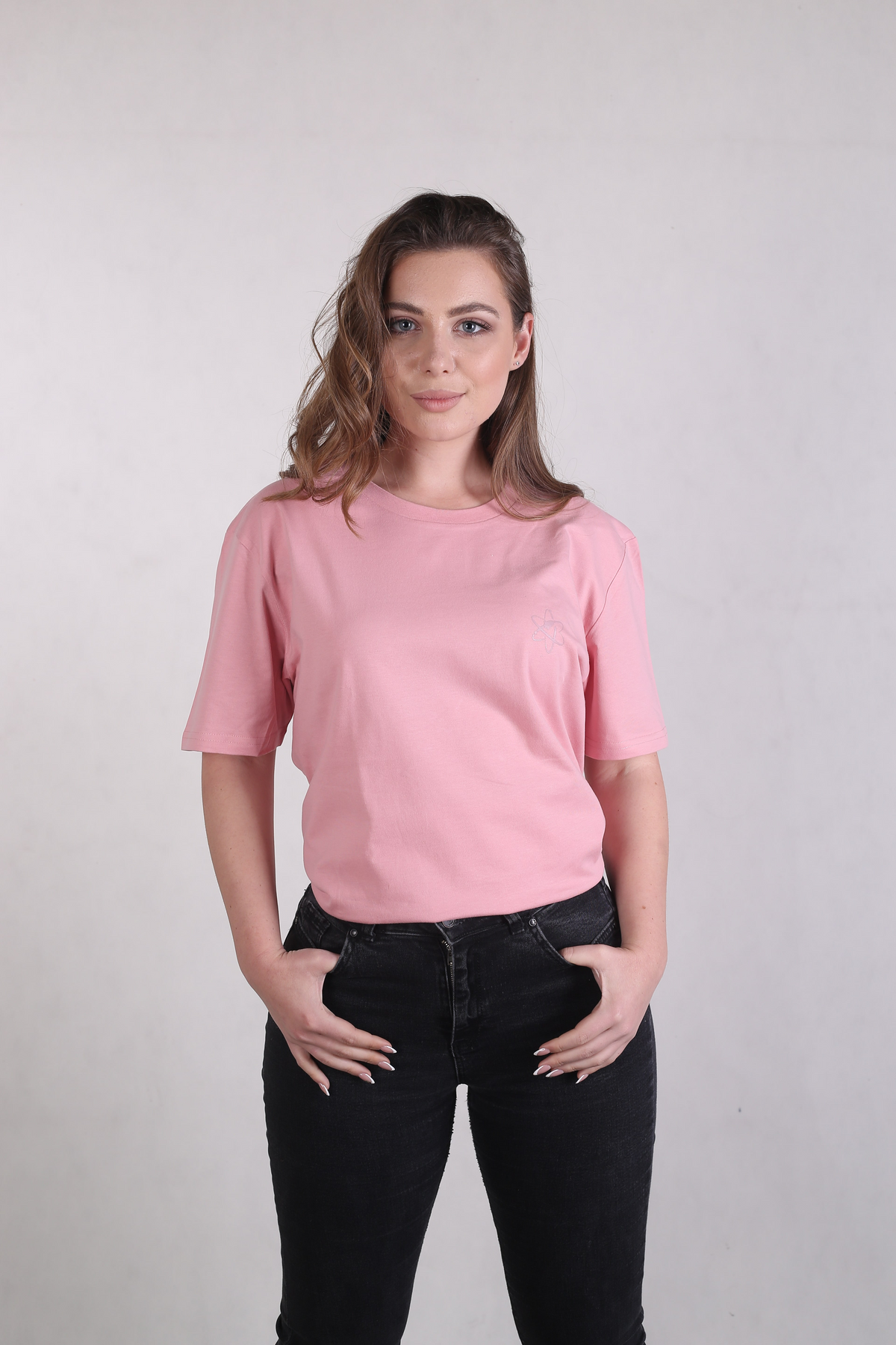 A woman modeling a light pink short-sleeve crewneck t-shirt with a small embroidered logo on the left chest, paired with black jeans, against a plain light gray background.