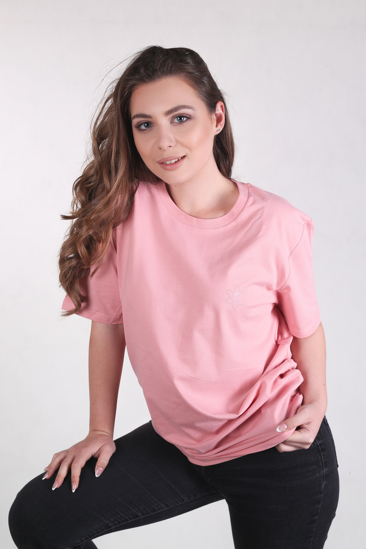 A woman wearing a light pink crewneck t-shirt and black jeans, posing against a light gray studio background.