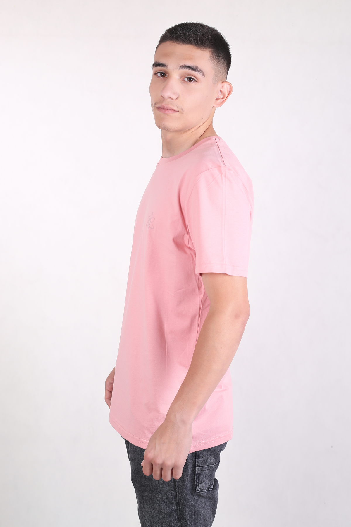 A man wearing a light pink crew-neck t-shirt and dark gray jeans, standing in profile against a plain white studio background.