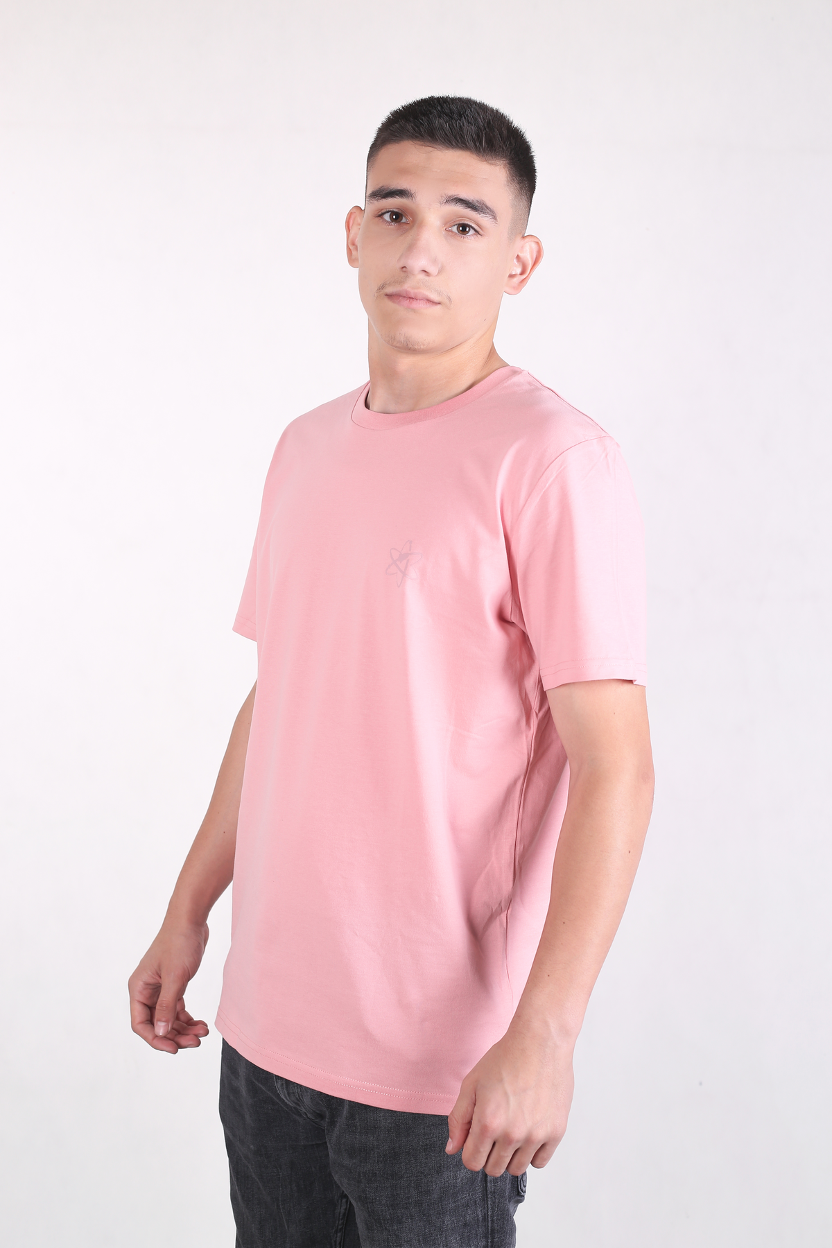 A young man wearing a light pink crewneck t-shirt and dark gray jeans, standing against a plain white studio background.