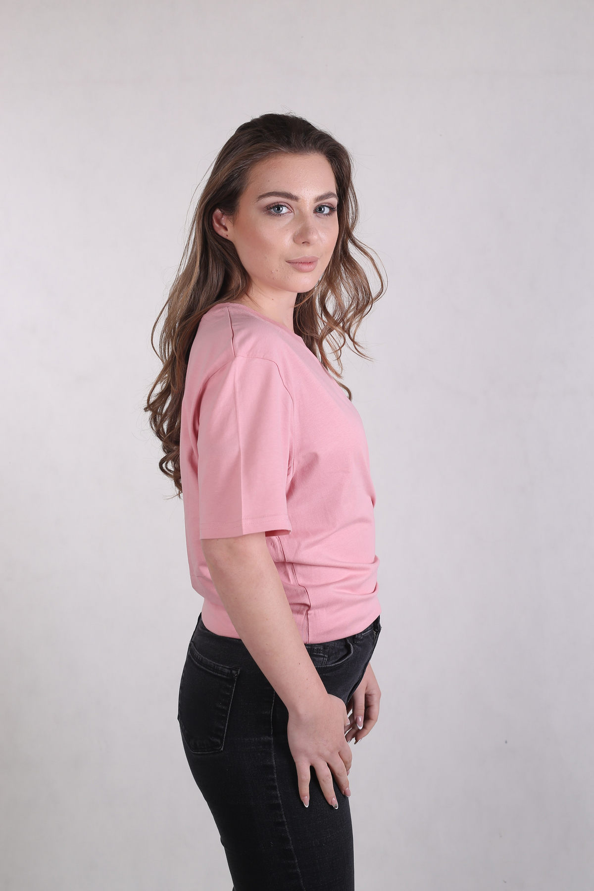 A woman with long brown hair is shown in profile wearing a light pink short-sleeve t-shirt and black jeans, standing against a plain light gray studio background.