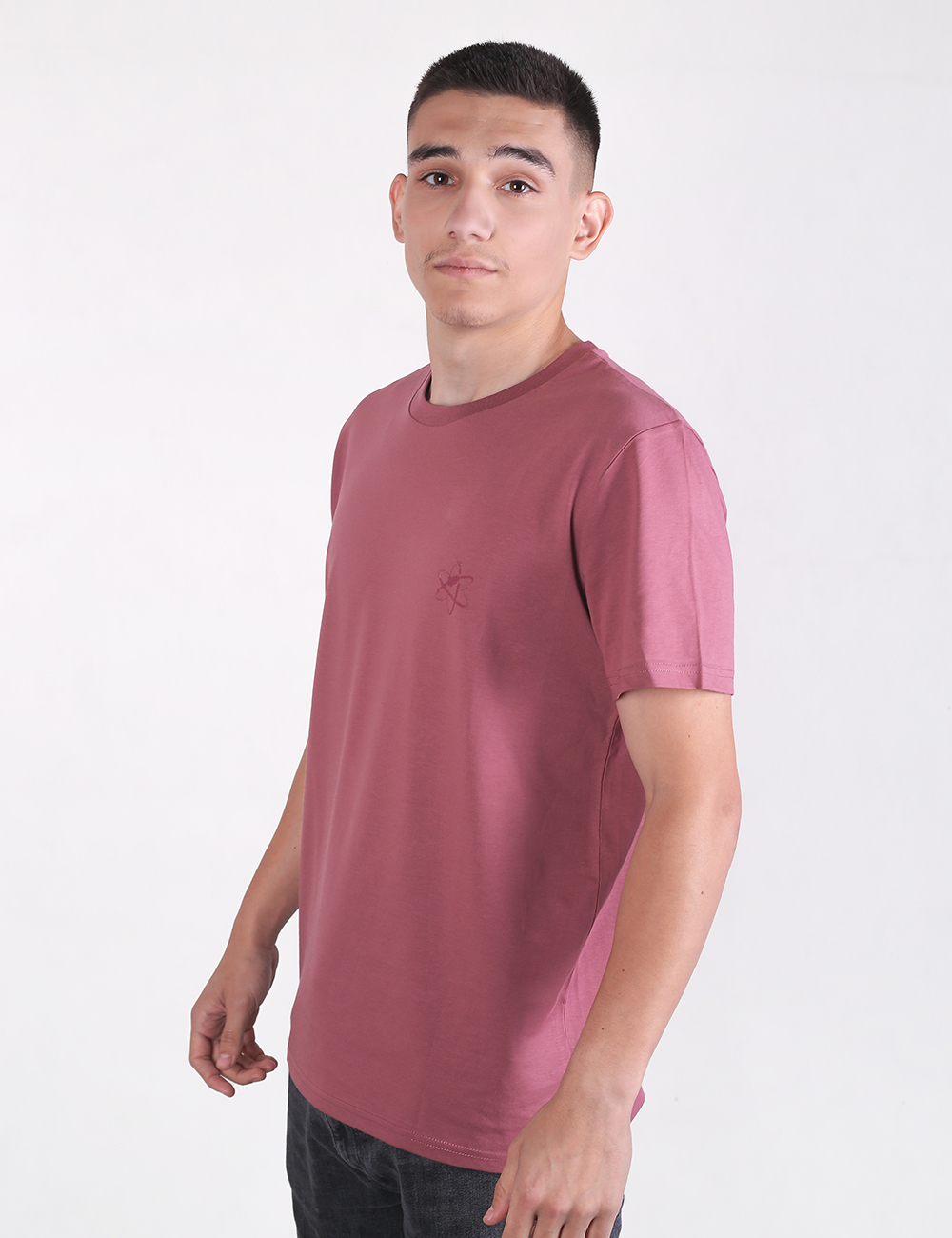 A young man with short dark hair standing in profile against a plain white background, wearing a mauve-pink crew neck t-shirt and dark pants.