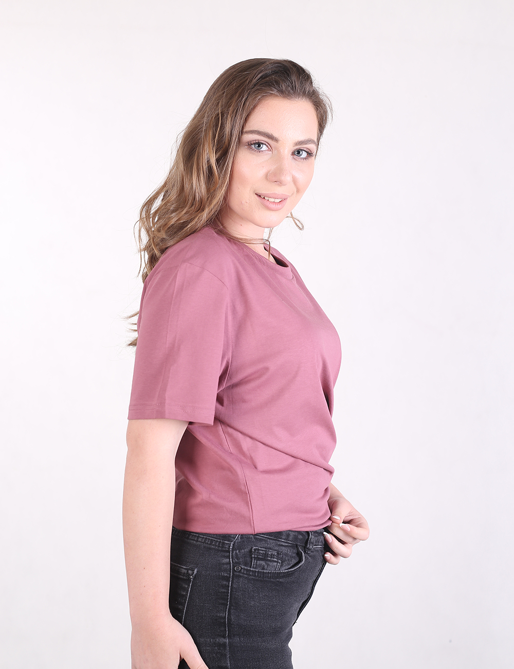 A side-profile shot of a woman with long light brown hair wearing a mauve short-sleeve t-shirt and black jeans. She is smiling at the camera against a plain white background.