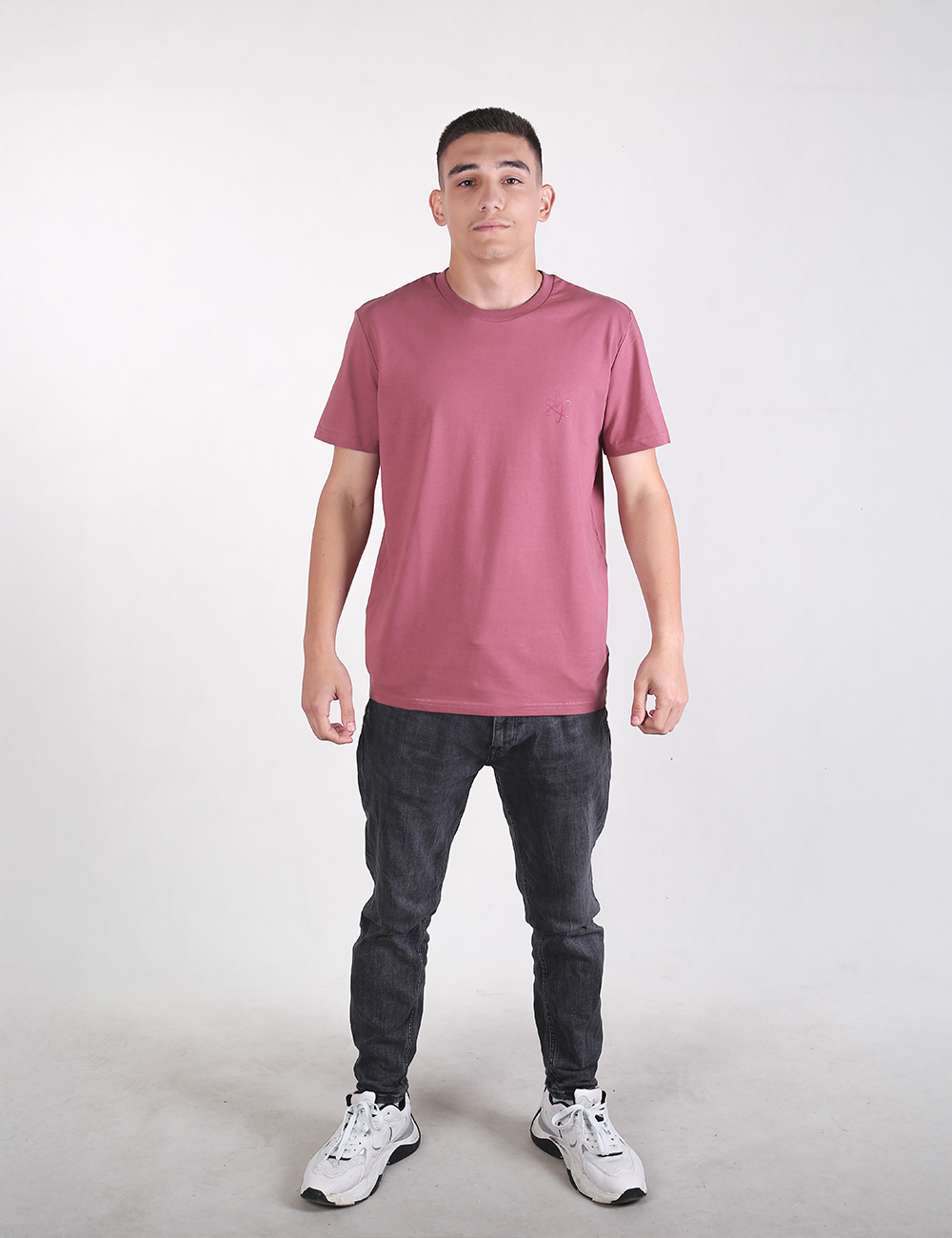 A young man stands facing forward against a plain light gray background. He is wearing a mauve, short-sleeved crew-neck t-shirt, dark charcoal or washed black slim-fit jeans, and white athletic sneakers with gray accents. His hair is cut short in a crew cut, and he has a neutral expression with his arms resting at his sides.