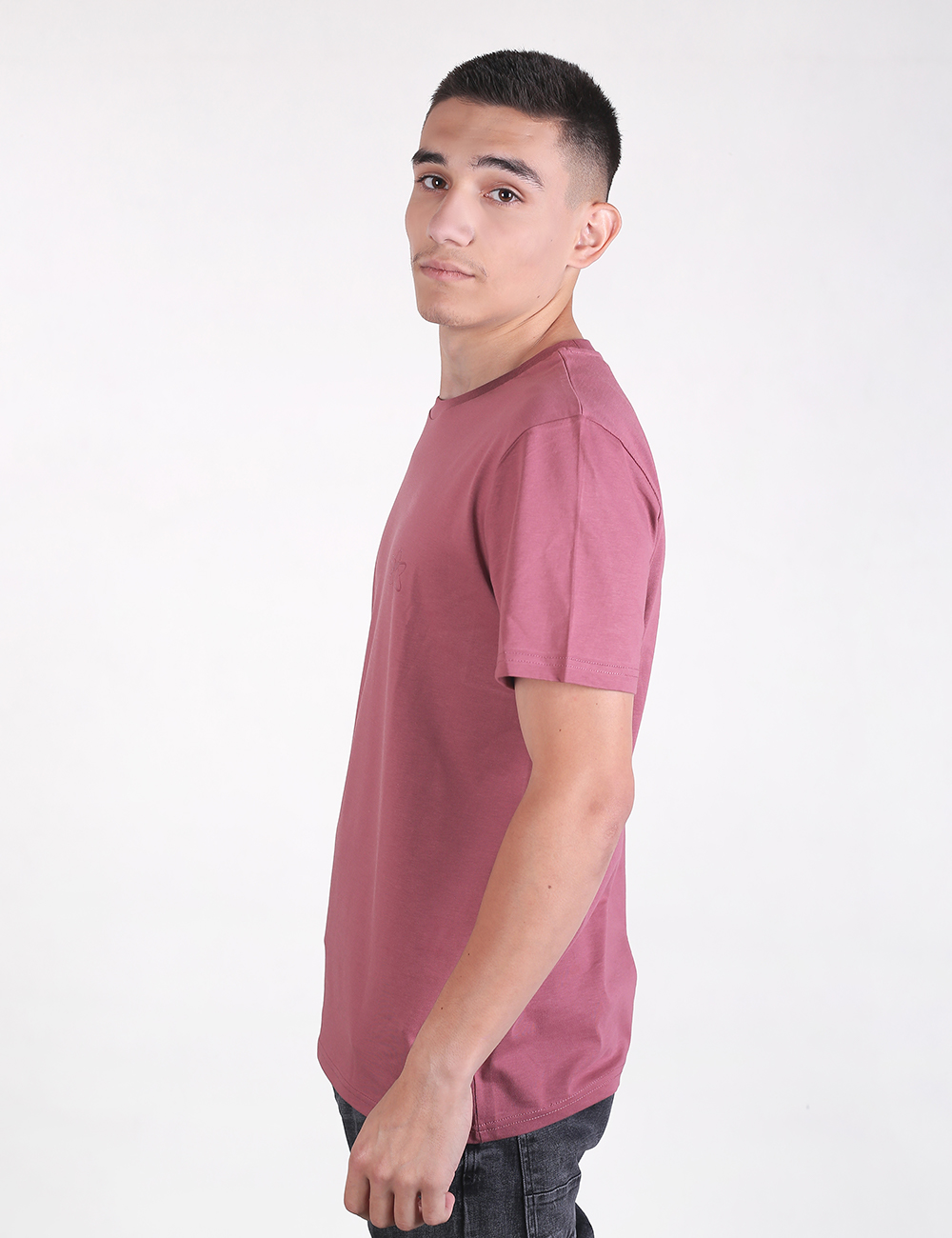 A young man with short dark hair, standing in a side-profile view against a plain, light grey background. He is wearing a solid mauve, short-sleeved crew neck t-shirt and dark grey or black jeans.