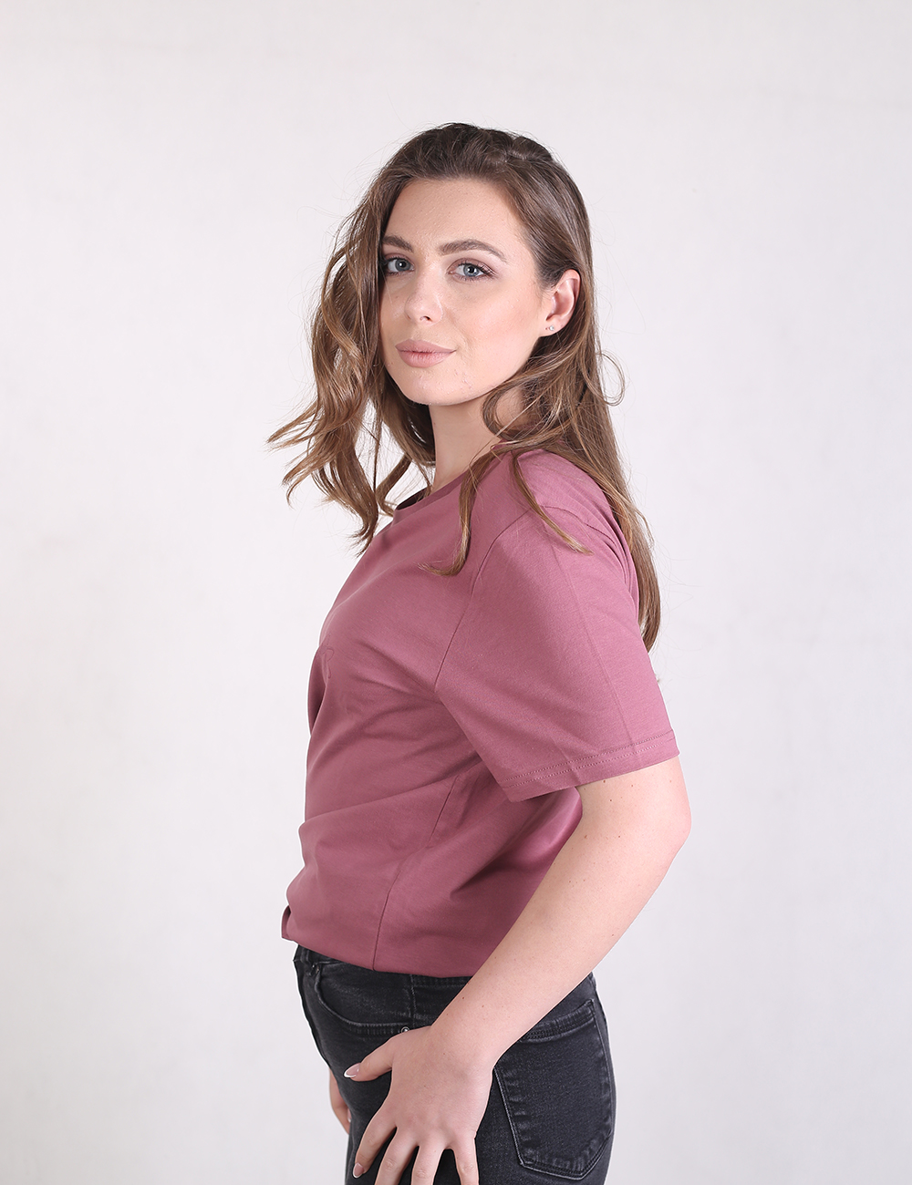 A woman with wavy brown hair looks toward the camera, wearing a solid mauve pink crew neck t-shirt tucked into black skinny jeans.